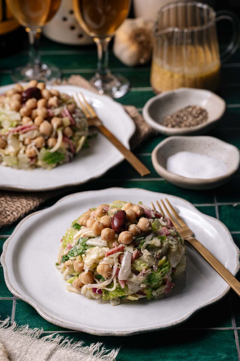 Two plates of La Scala salad with the signature olive on top, chickpeas, radishes, and greens on white dishes, set on a green tile table with cutlery, condiments, and drinks in the background.