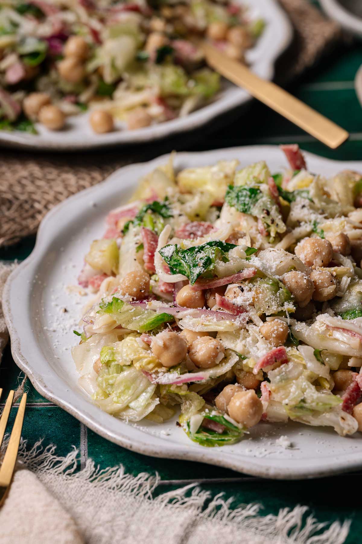A plate of Italian chopped salad with lettuce, chickpeas, sliced red onions, salami, and grated cheese on a white plate with a gold fork beside it.