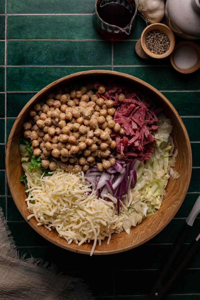 A wooden bowl contains chickpeas, sliced salami, shredded lettuce, shredded cheese, and chopped red onions, arranged in sections on a green tiled surface.