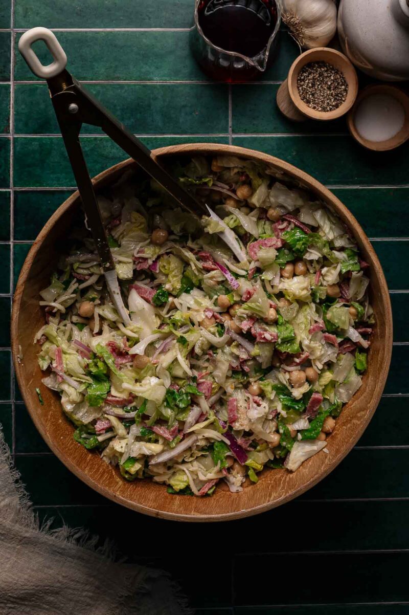 A wooden bowl of La Scala chopped salad with lettuce, chickpeas, and other vegetables on a dark green tiled surface, with salad tongs and seasonings nearby.