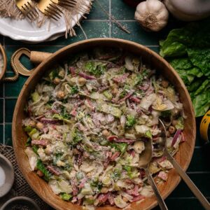 A large wooden bowl filled with La Scala chopped salad topped with grated cheese, surrounded by serving utensils, garlic, lettuce, a glass of red wine, and plates with forks.