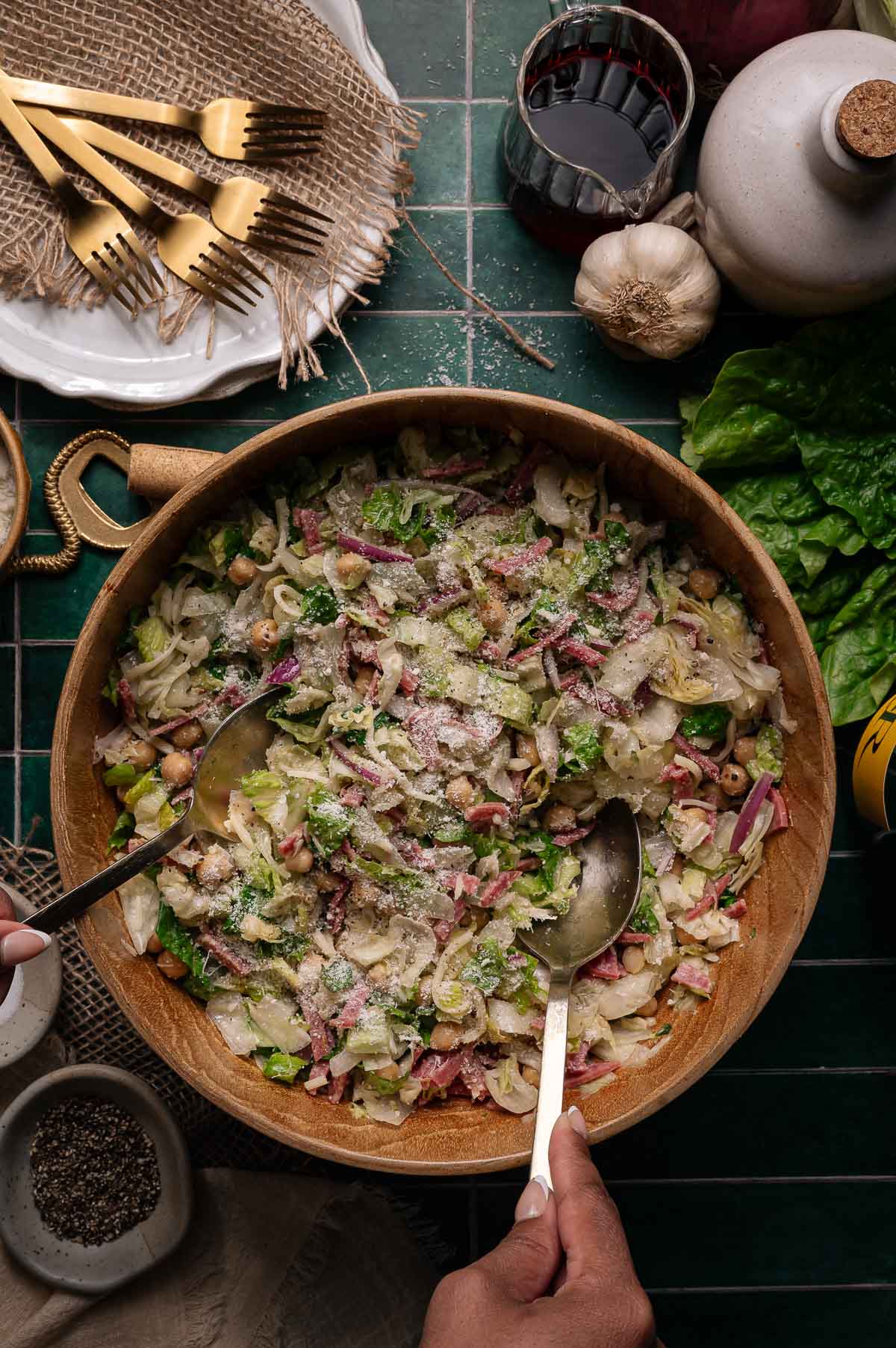 A large wooden bowl filled with a Italian chopped salad topped with grated cheese, surrounded by salad utensils, wine, garlic, lettuce, and tableware on a green tiled surface.