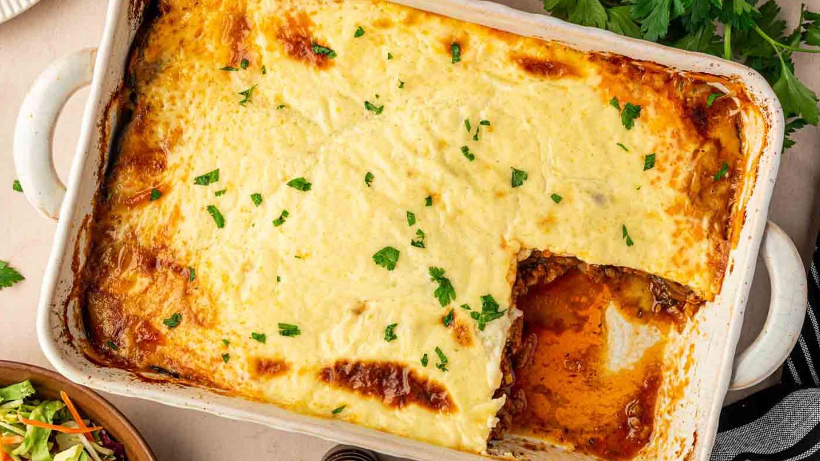 A baked casserole dish of moussaka topped with a golden-brown cheese layer, garnished with chopped parsley, with one serving removed from the corner.