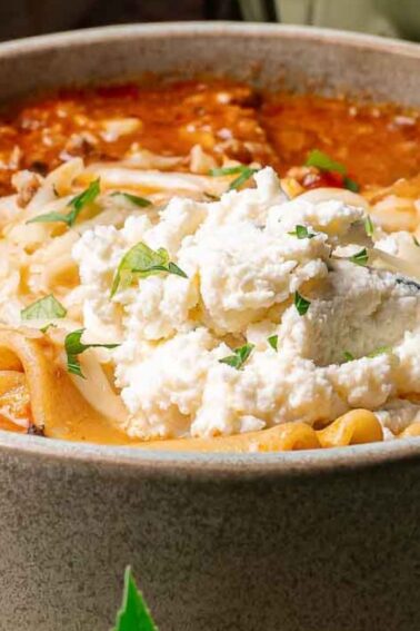 A bowl of lasagna soup topped with ricotta cheese, shredded cheese, and fresh herbs, with a spoon inside the bowl.