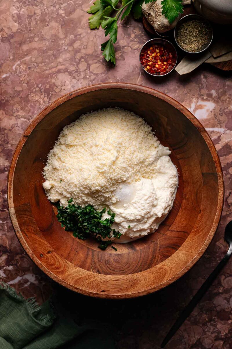 A wooden bowl containing ricotta cheese, grated parmesan, and chopped herbs on a marble countertop, with seasonings and parsley nearby.