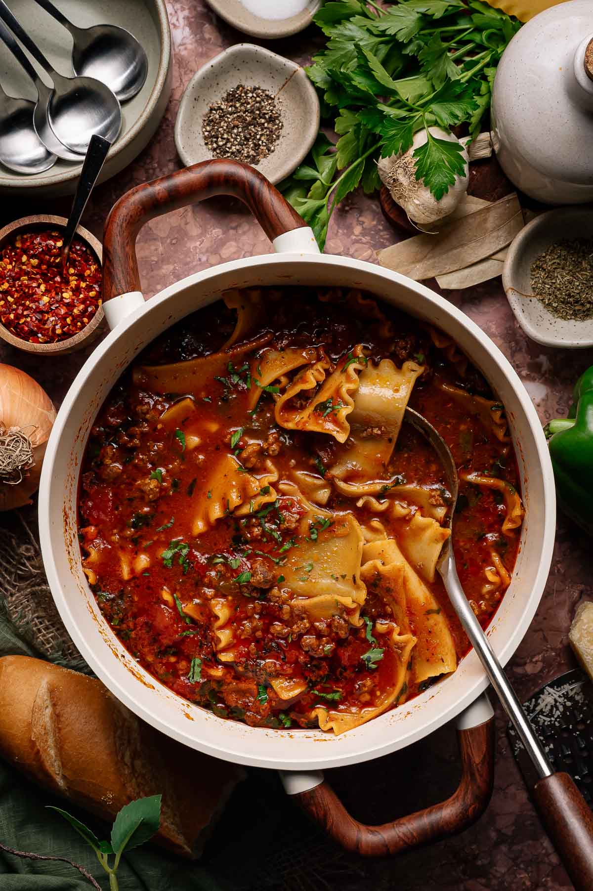A pot of lasagna soup with wide noodles, ground meat, tomato broth, and herbs sits on a countertop surrounded by spices, bread, parsley, and cooking utensils.