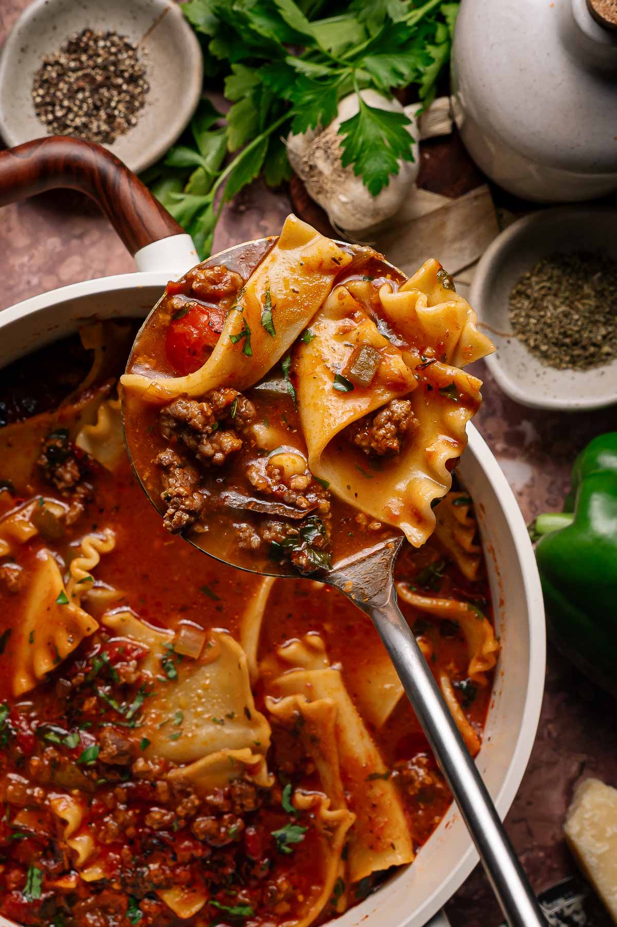 A ladle scoops up lasagna soup with ground meat, pasta, tomatoes, and herbs from a pot. Surrounding the pot are spices, herbs, and vegetables.