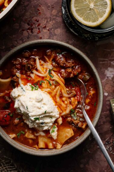 A bowl of lasagna soup topped with shredded cheese and a dollop of ricotta, with a spoon in the bowl and lemon slices on a plate nearby.