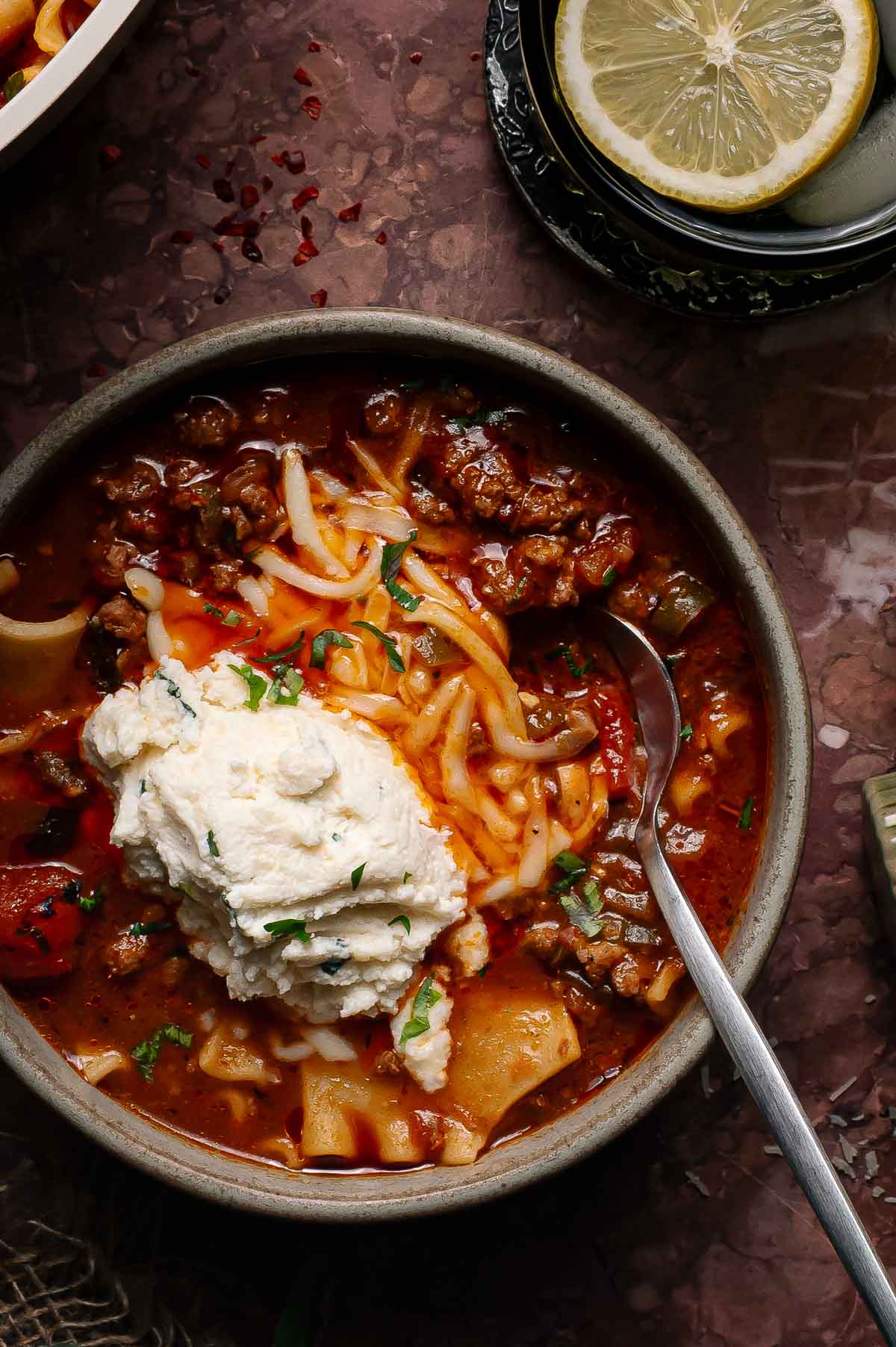 A bowl of lasagna soup topped with shredded cheese and a dollop of ricotta, with a spoon in the bowl and lemon slices on a plate nearby.
