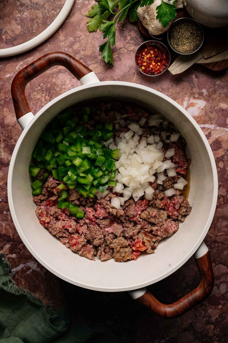 A pot contains partially cooked ground beef and sausage, chopped green bell pepper, and diced onion on a brown marble countertop, with seasonings and herbs nearby.