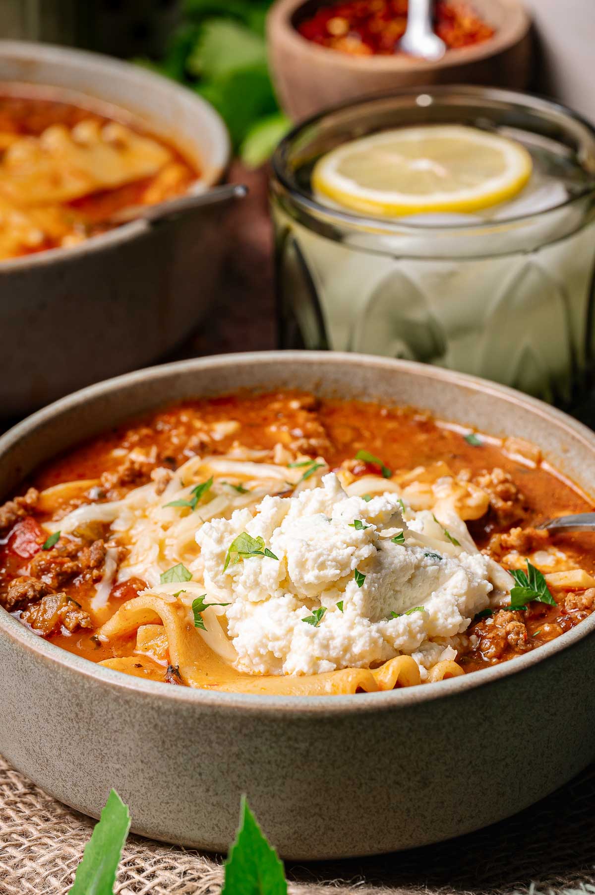 A bowl of lasagna soup topped with ricotta cheese and herbs, with a glass of water and a slice of lemon in the background.