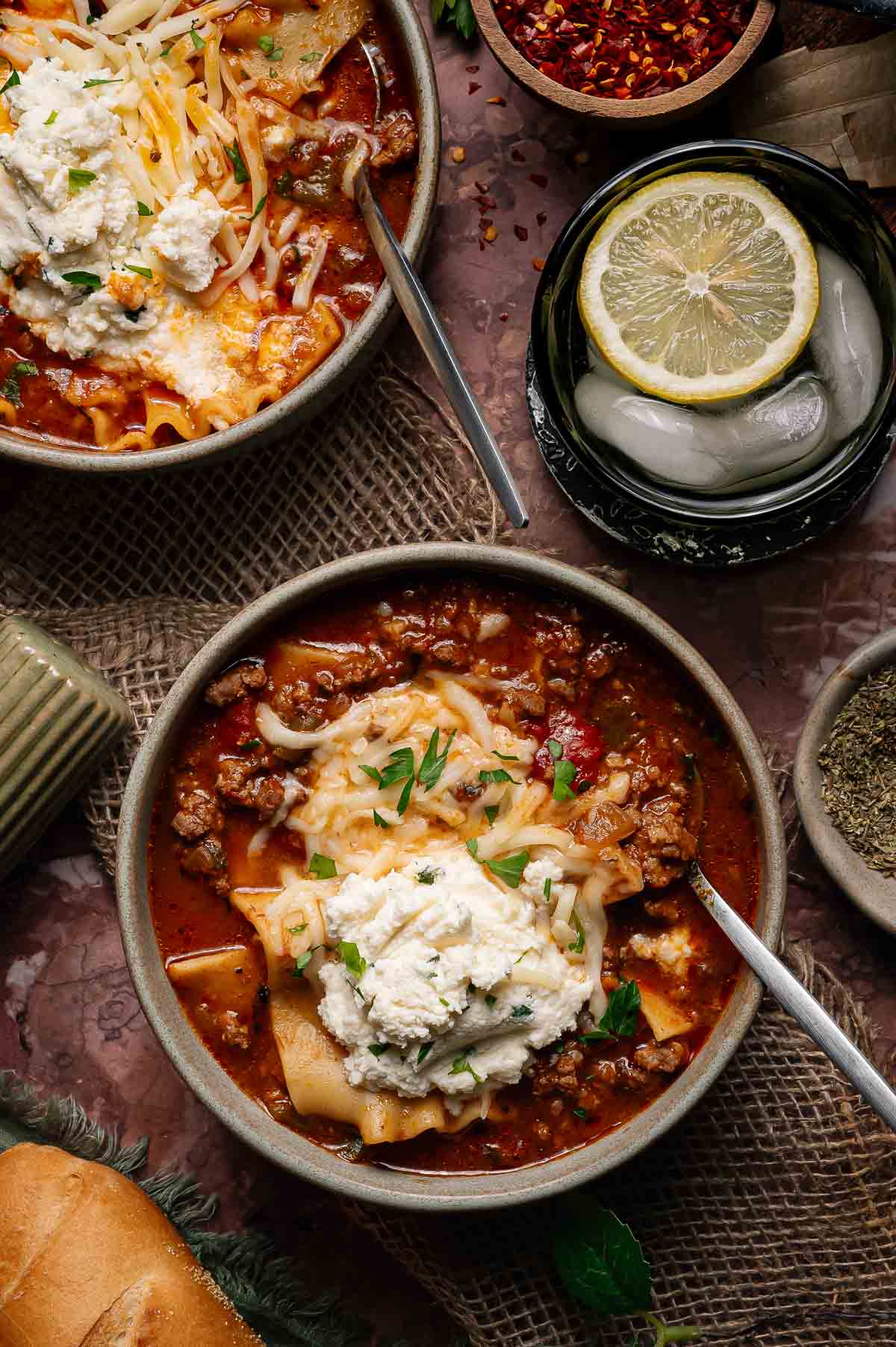 Two bowls of lasagna soup topped with shredded cheese and ricotta, surrounded by ingredients and a drink with a lemon slice and ice.