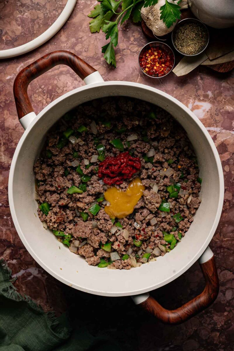 Ground beef and sausage, diced onion, and green pepper cooking in a white pot, topped with tomato paste and mustard. Surrounding the pot are herbs and small bowls of spices.