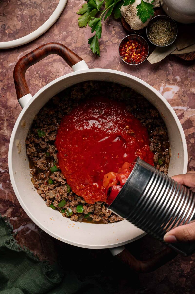 A hand pours canned tomatos into a white pot with browned ground beef and chopped vegetables on a brown countertop.