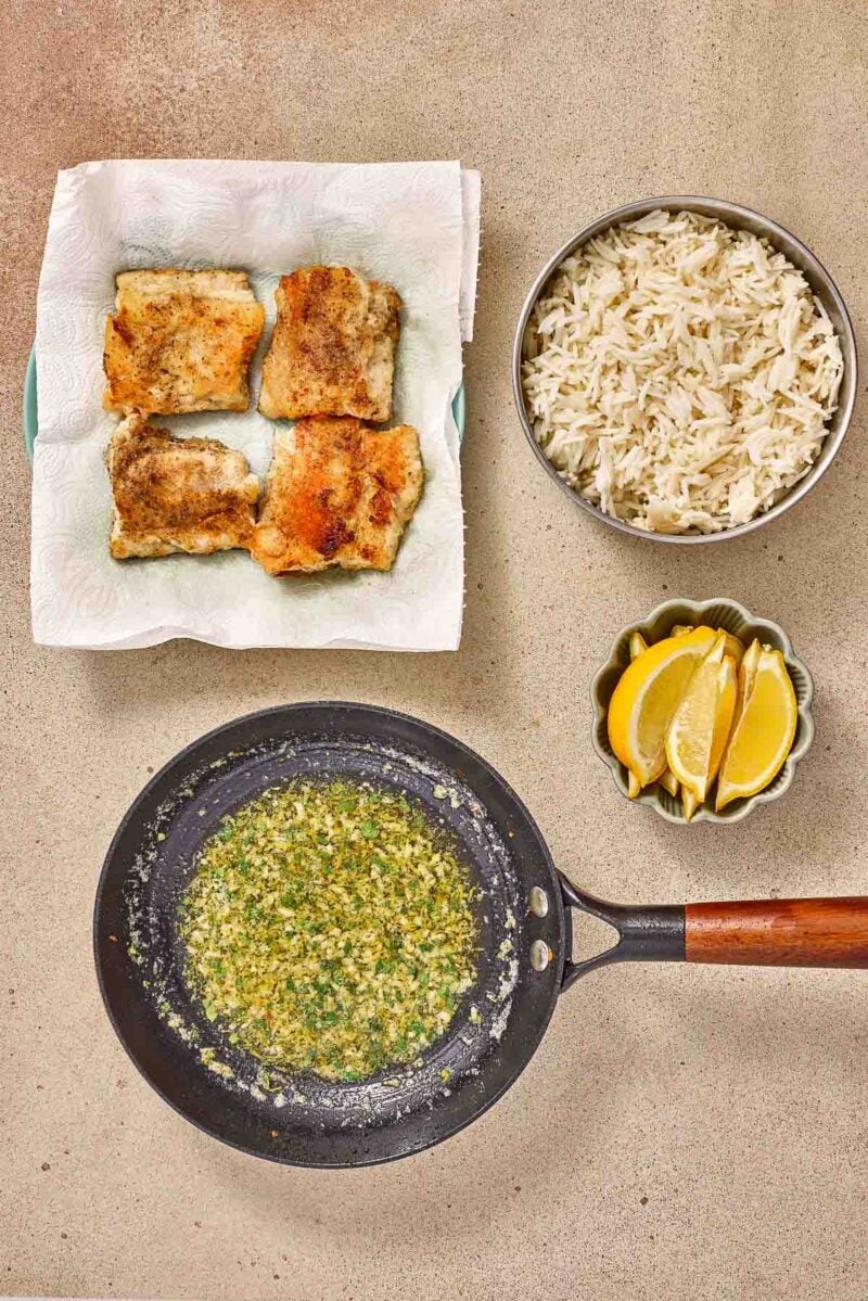 Four pieces of cooked fish on a paper towel, a bowl of white rice, lemon wedges, and a skillet with garlic lemon butter sauce on a beige surface.