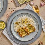 A plate of white rice and cod with a garlic lemon butter sauce garnished with lemon slices, served on a gray plate with gold cutlery, surrounded by lemon, garlic, and peppercorns.