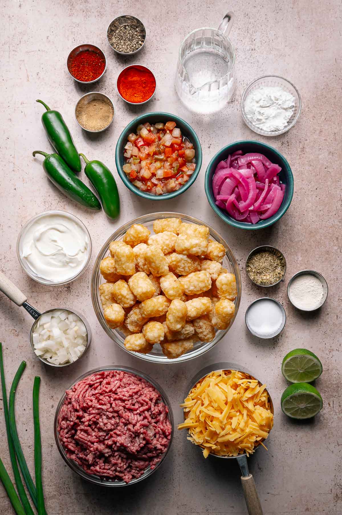 Ingredients for a loaded totchos laid out on a counter, including ground beef, cheese, tater tots, jalapeños, spices, sour cream, limes, and various toppings.