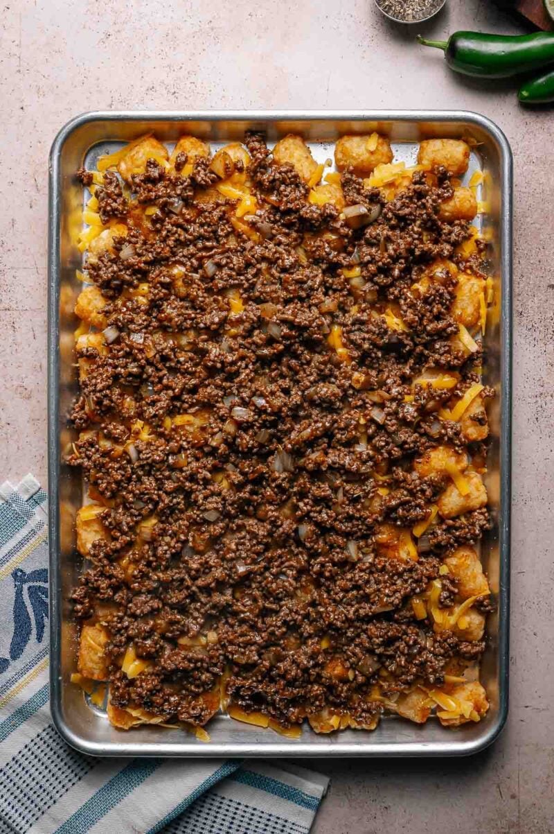 A baking sheet topped with tater tots, melted cheese, ground beef, and onions, arranged on a kitchen towel next to whole jalapeños and a pepper shaker.