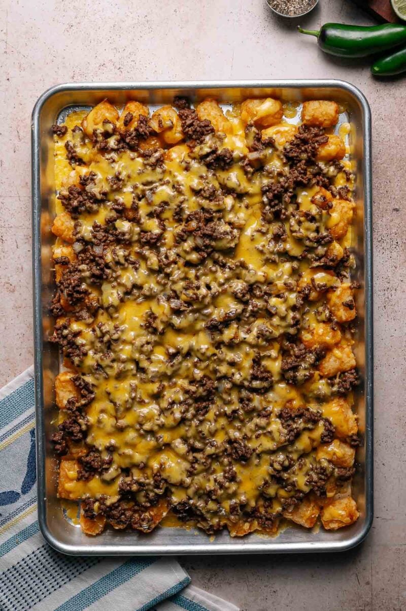 Rectangular baking tray filled with tater tots topped with cooked ground beef and melted cheese, photographed from above on a light-colored surface.