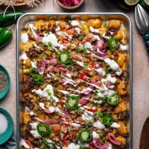 A sheet pan of loaded tater tots topped with ground beef, melted cheese, jalapeños, pickled onions, pico de gallo, green onions, and drizzled with sour cream.