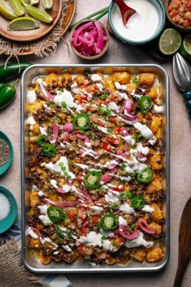 A sheet pan of loaded tater tots topped with ground beef, melted cheese, jalapeños, pickled onions, pico de gallo, green onions, and drizzled with sour cream.
