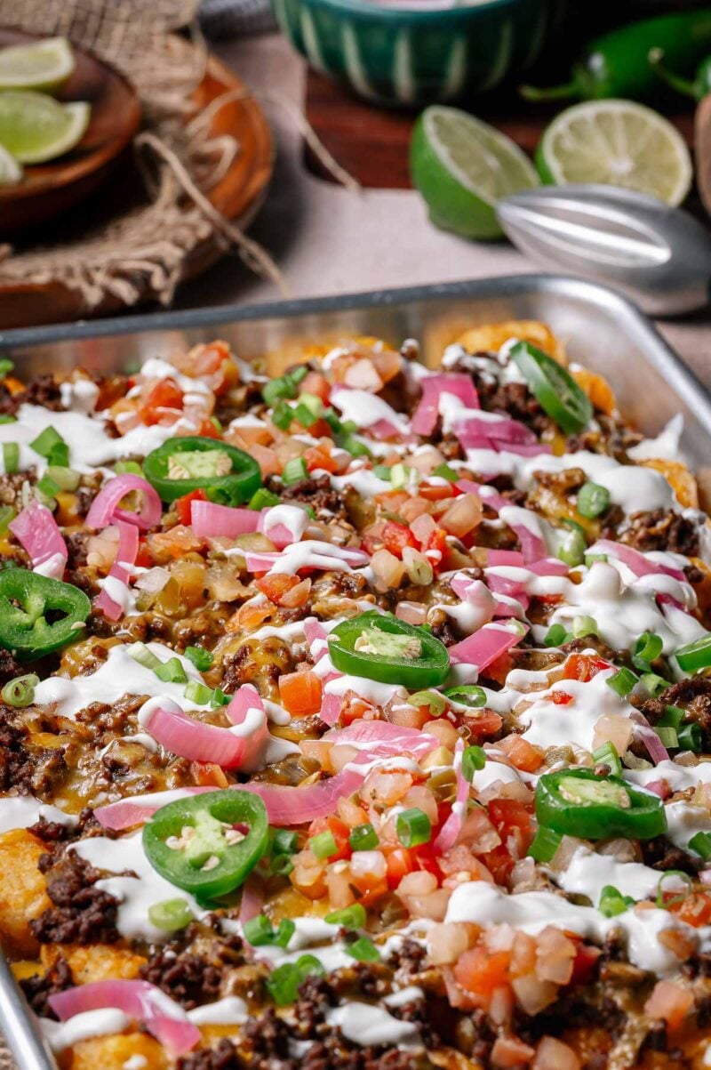 A baking sheet filled with loaded tater tots topped with ground beef, sliced jalapeños, diced tomatoes, pickled onions, green onions, and drizzled with white sauce.