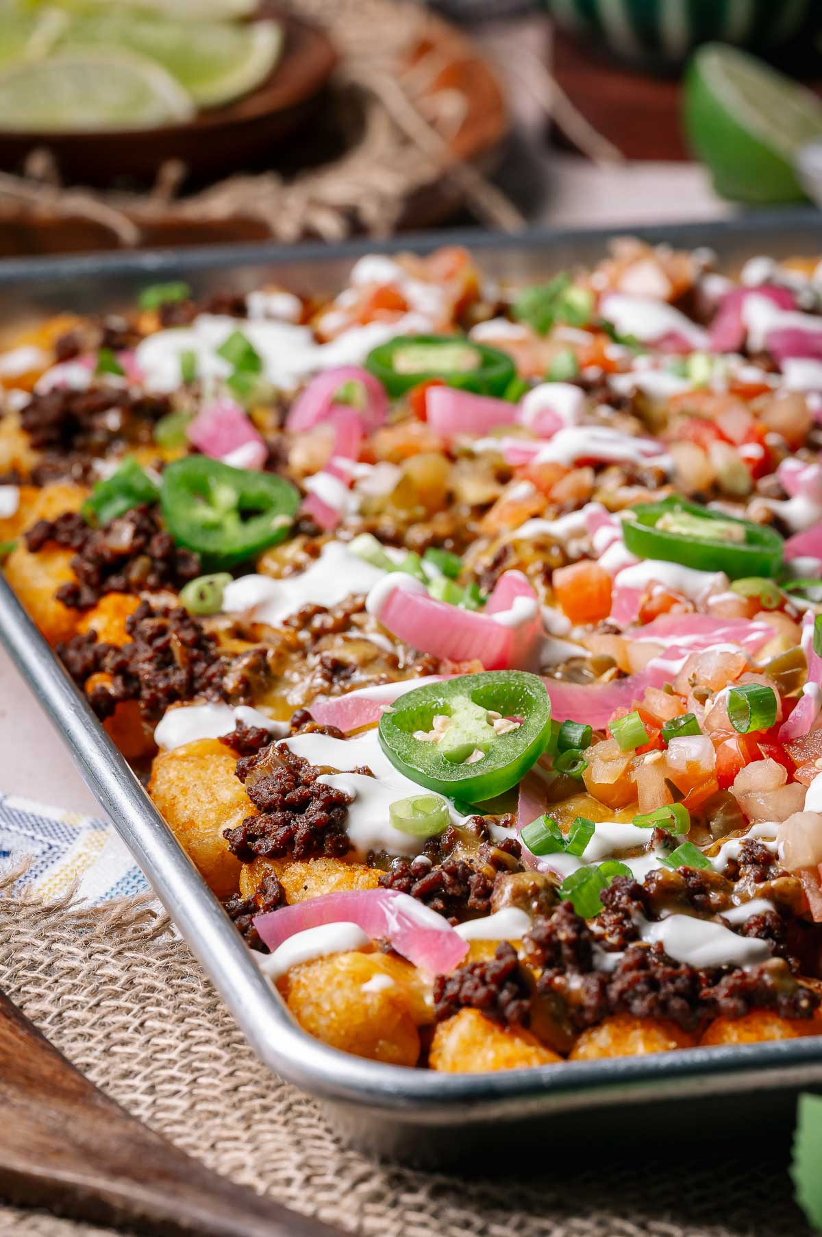 Sheet pan loaded tater tots topped with ground beef, melted cheese, jalapeño slices, diced tomatoes, green onions, pickled onions, and drizzled with sour cream.