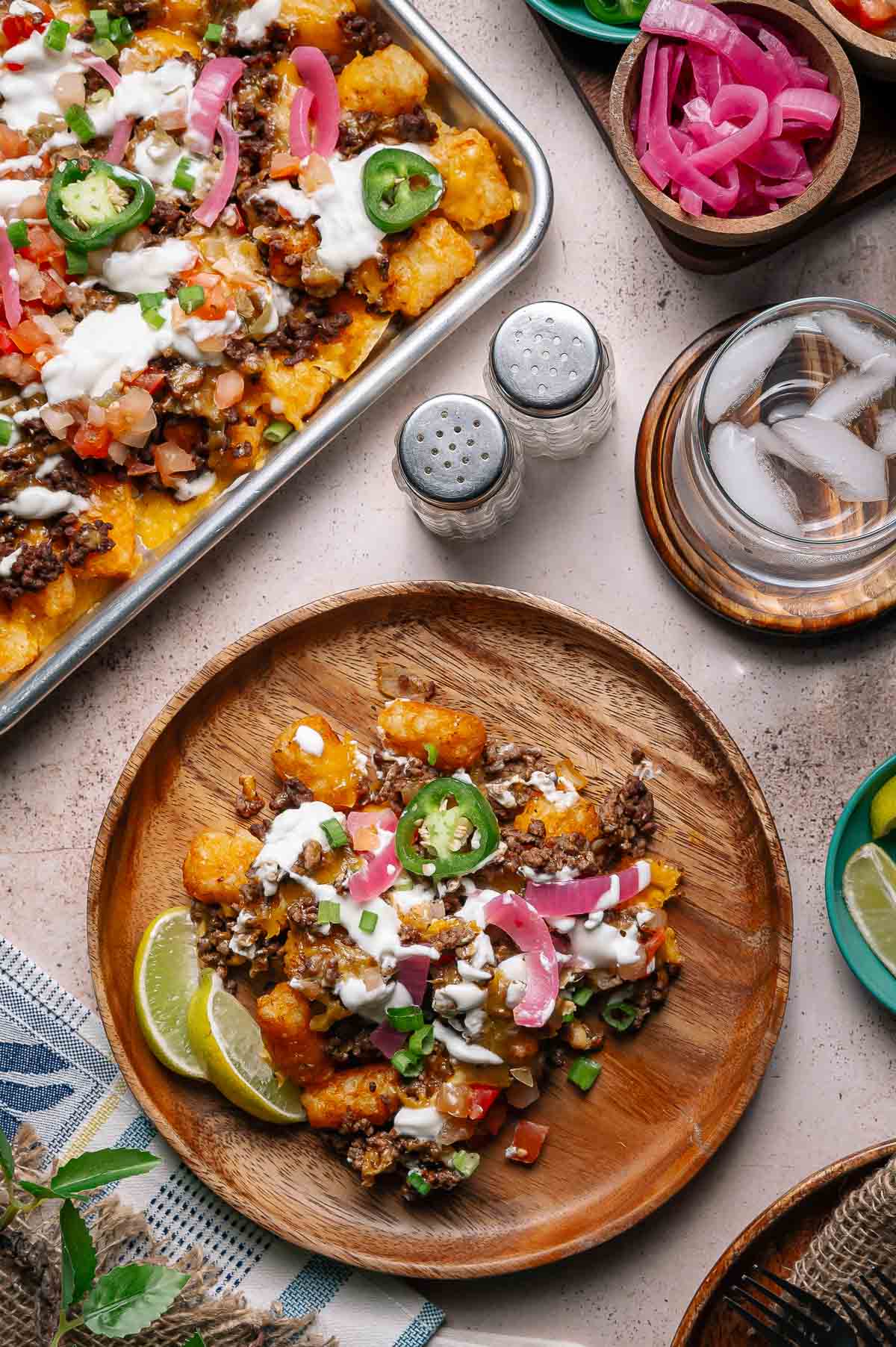 A wooden plate with loaded tater tots topped with ground beef, cheese, jalapeños, pickled onions, sour cream, and lime wedges; a tray of the same dish and condiments are nearby.