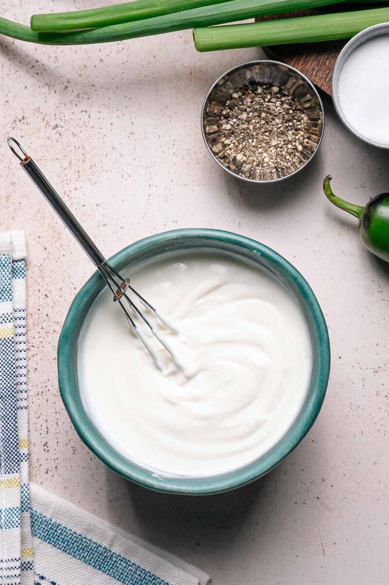 A bowl of lime crema white sauce with a whisk inside, next to a towel, chopped green onions, a jalapeño, ground pepper, and a bowl of salt on a light surface.