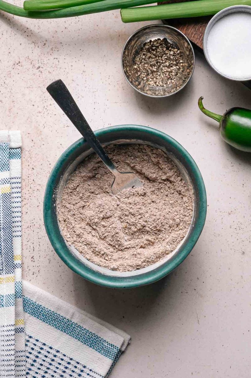 A bowl filled with a dry taco seasoning mixture and a fork, surrounded by a jalapeño, a bowl of pepper, green onions, and a kitchen towel on a countertop.