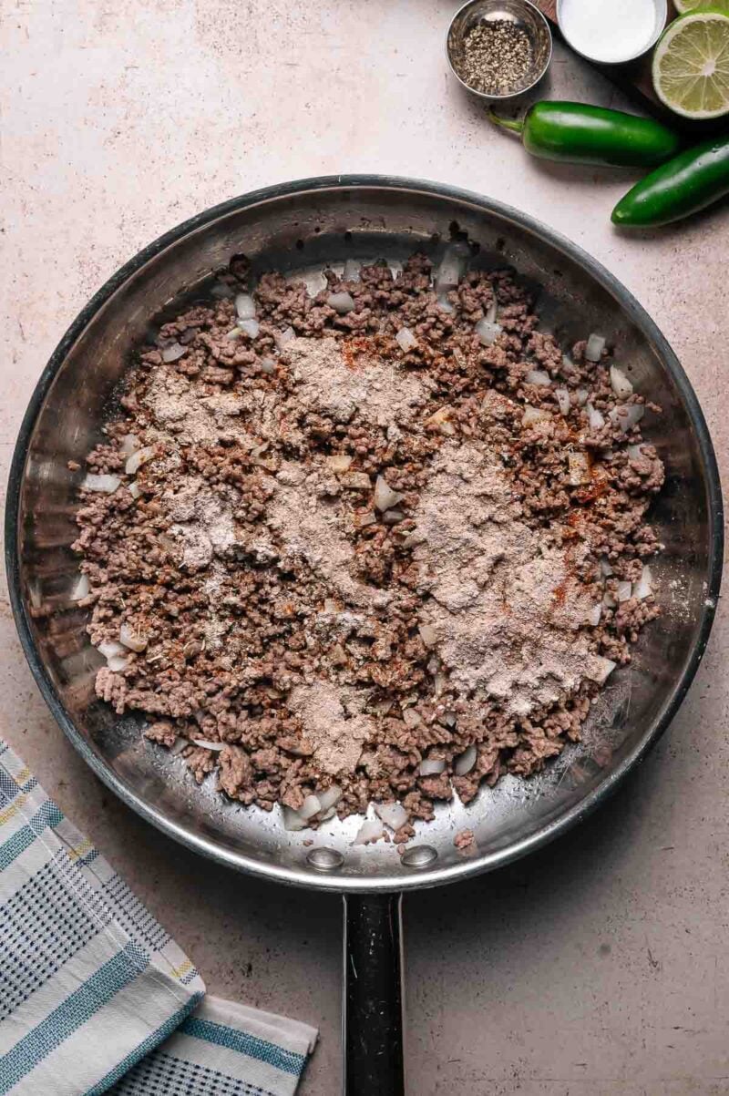 A skillet with cooked ground beef and chopped onions, topped with dry seasoning mix. Nearby are a towel, lime, jalapeños, salt, and pepper.