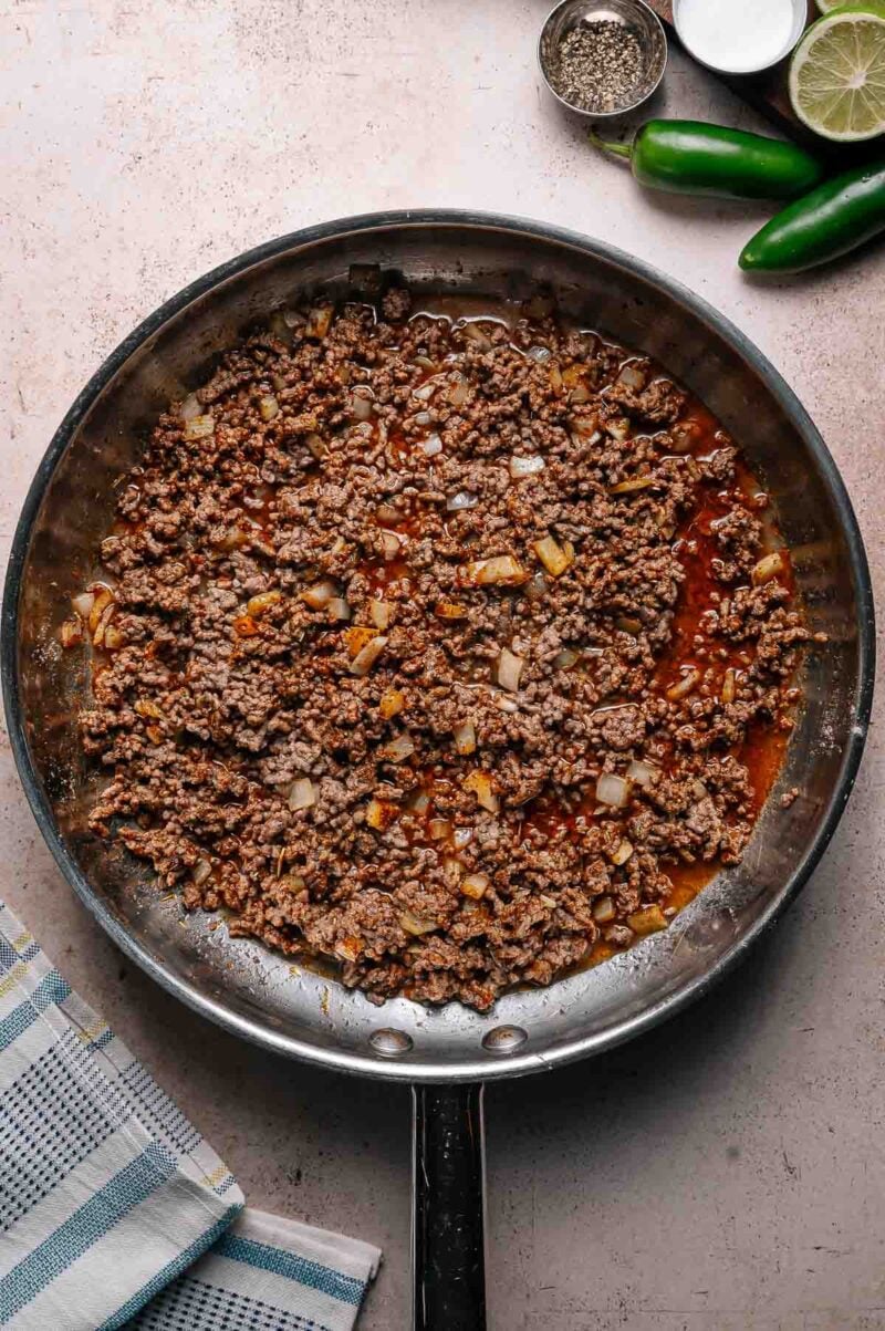 A skillet filled with cooked ground beef mixed with diced onions and spices, placed on a light countertop next to lime, jalapeño, and small bowls of seasoning.