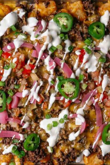 A sheet pan topped with tater tots, ground beef, melted cheese, sliced jalapeรฑos, diced tomatoes, pickled onions, green onions, and drizzled with white sauce.