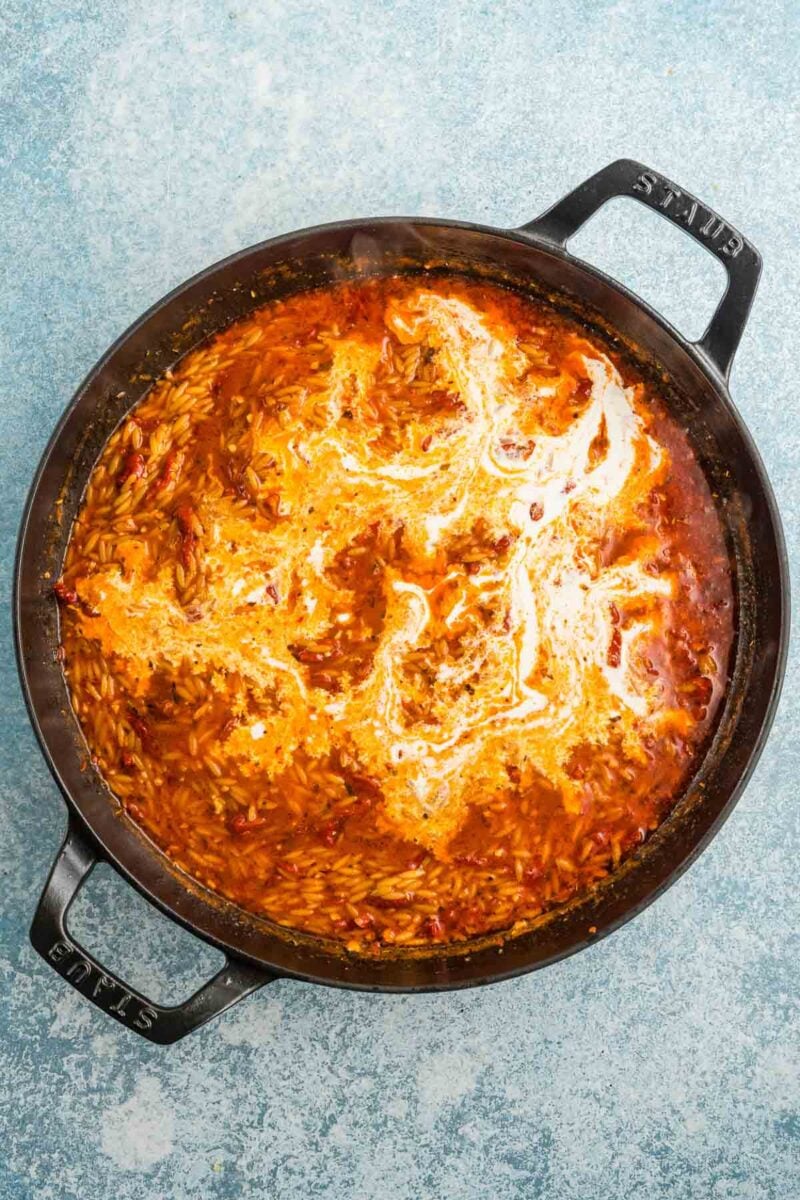 Large black pot containing tomato-based orzo pasta with a swirl of cream on top, set on a light blue textured surface.