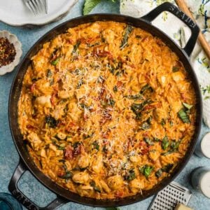 A skillet filled with creamy marry me chicken orzo mixed with chicken, basil, sun-dried tomatoes, and grated cheese is surrounded by plates, utensils, and fresh basil.