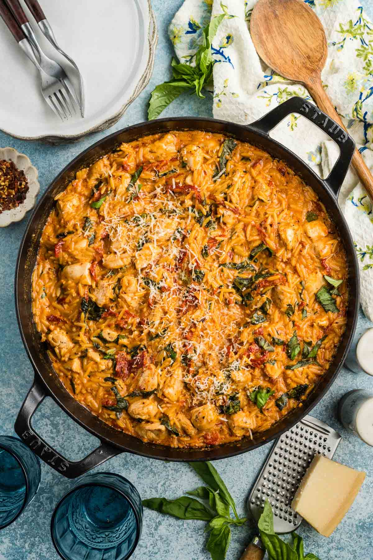 A skillet filled with creamy marry me chicken orzo mixed with chicken, basil, sun-dried tomatoes, and grated cheese is surrounded by plates, utensils, and fresh basil.