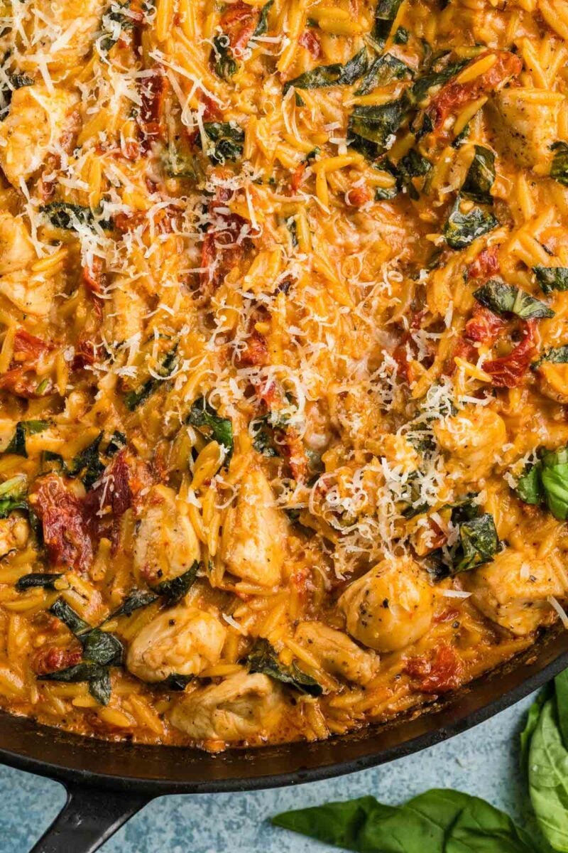 Skillet filled with cooked orzo pasta, chunks of chicken, basil, sun-dried tomatoes, and grated cheese, garnished with fresh basil.