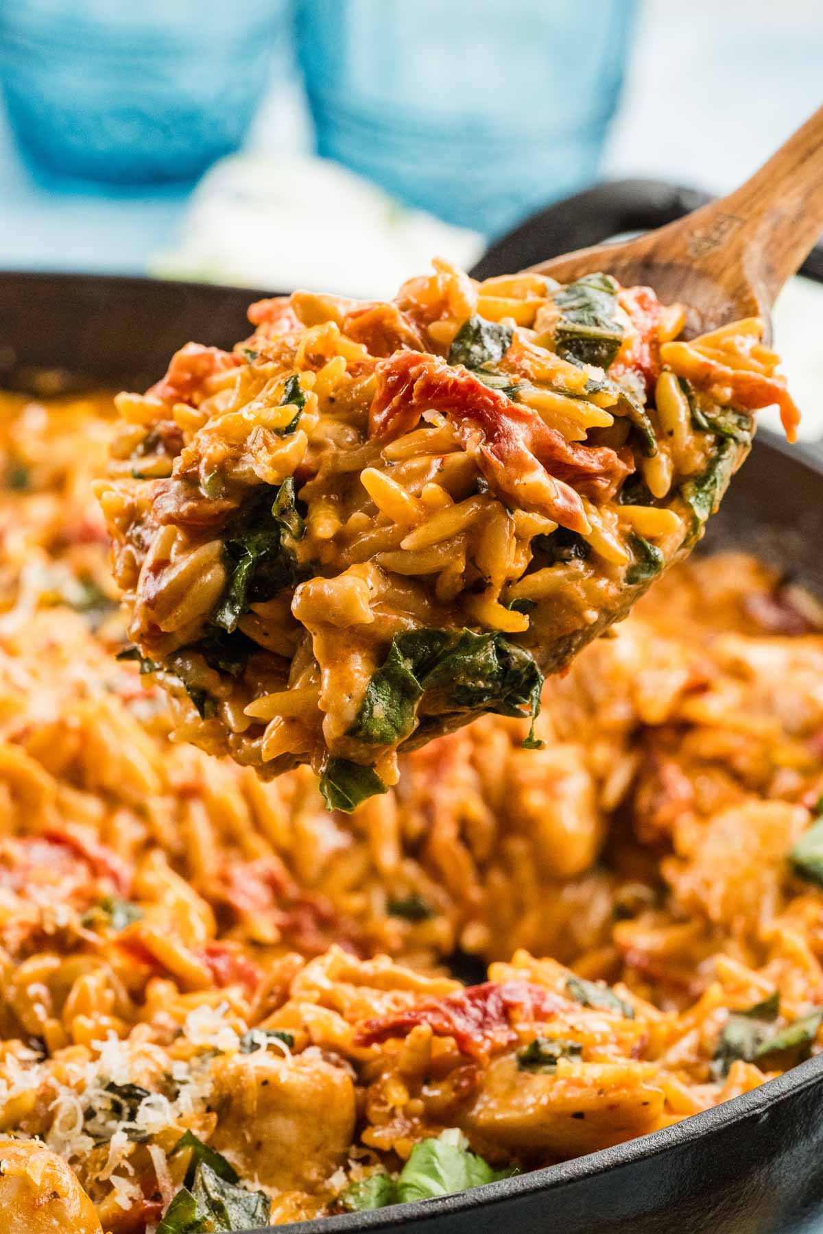 A wooden spoon lifts a serving of creamy marry me chicken orzo with basil, sun-dried tomatoes, and chicken from a skillet.