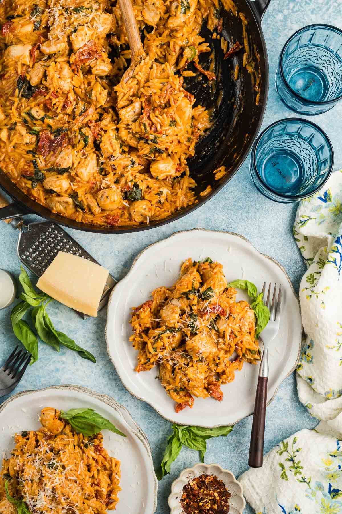 A skillet and plates of creamy marry me chicken orzo with chicken, spinach, and sun-dried tomatoes, garnished with grated cheese and basil, next to glasses of water and a block of cheese.
