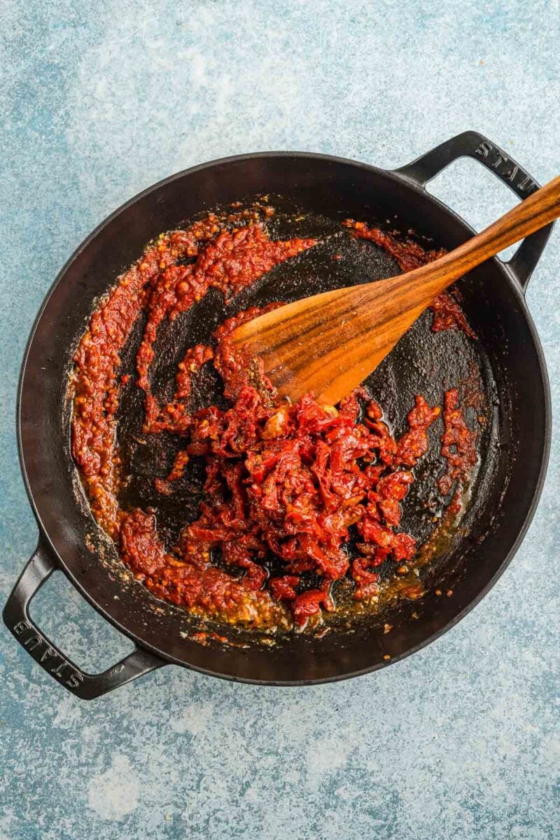 A cast iron skillet contains cooked tomato paste being stirred with a wooden spatula on a blue textured surface.