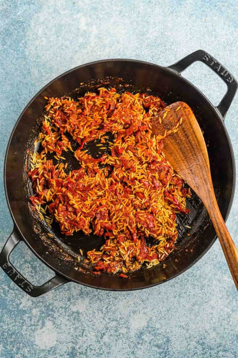 A cast iron skillet containing uncooked orzo mixed with tomato sauce and seasonings, with a wooden spatula resting inside.