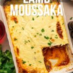 A baked dish of lamb moussaka topped with creamy sauce, with a serving removed to show the layers. Parsley garnish and kitchen utensils are nearby. Text overlay presents a recipe.