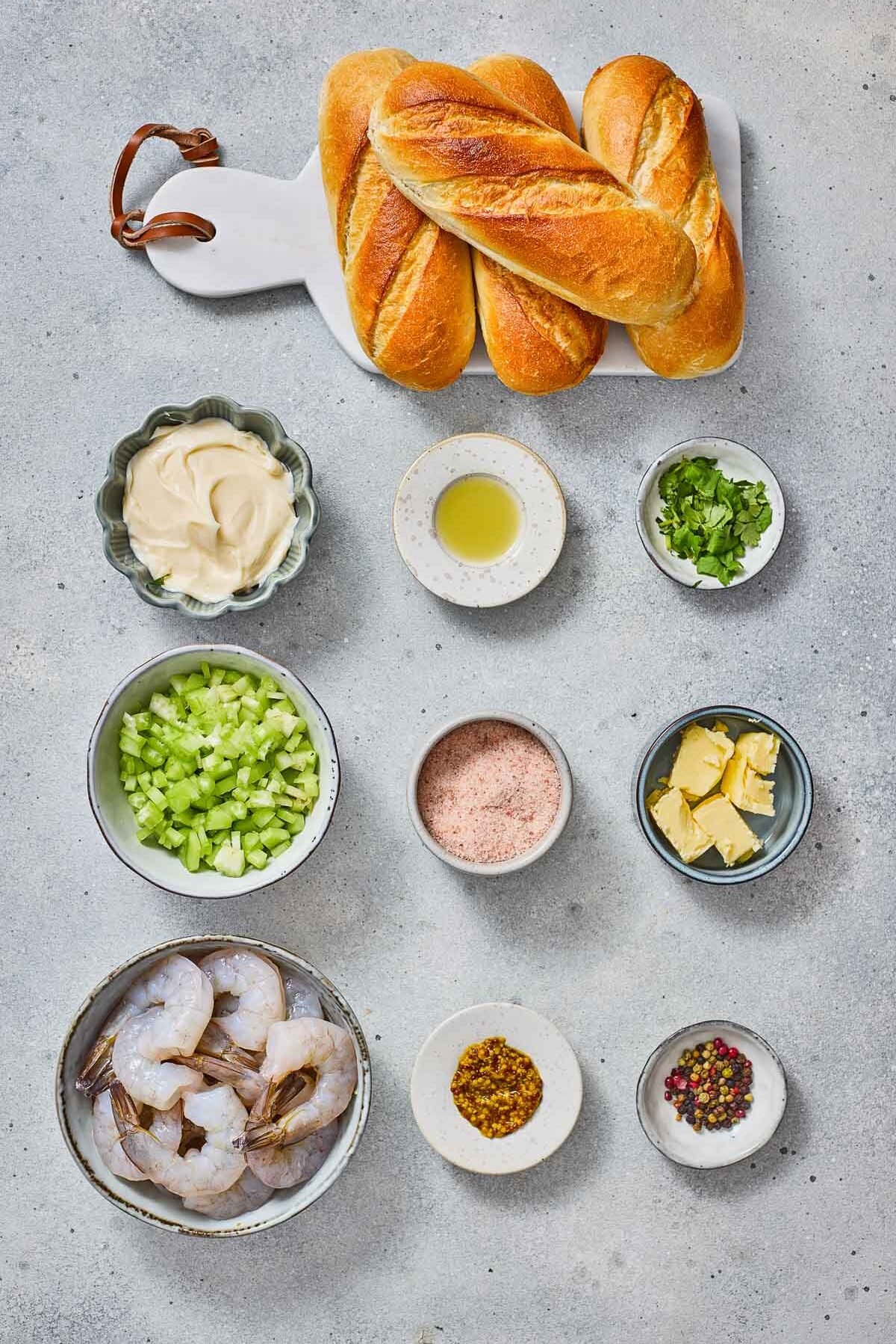 Assorted ingredients for a shrimp roll: baguettes, shrimp, celery, mayonnaise, oil, butter, herbs, spices, mustard, and salt arranged on a light surface.
