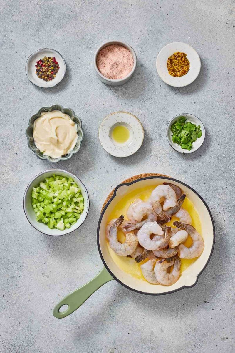 A skillet with raw shrimp in melted butter, surrounded by bowls of chopped celery, mayonnaise, spices, herbs, and lemon juice on a light gray surface.