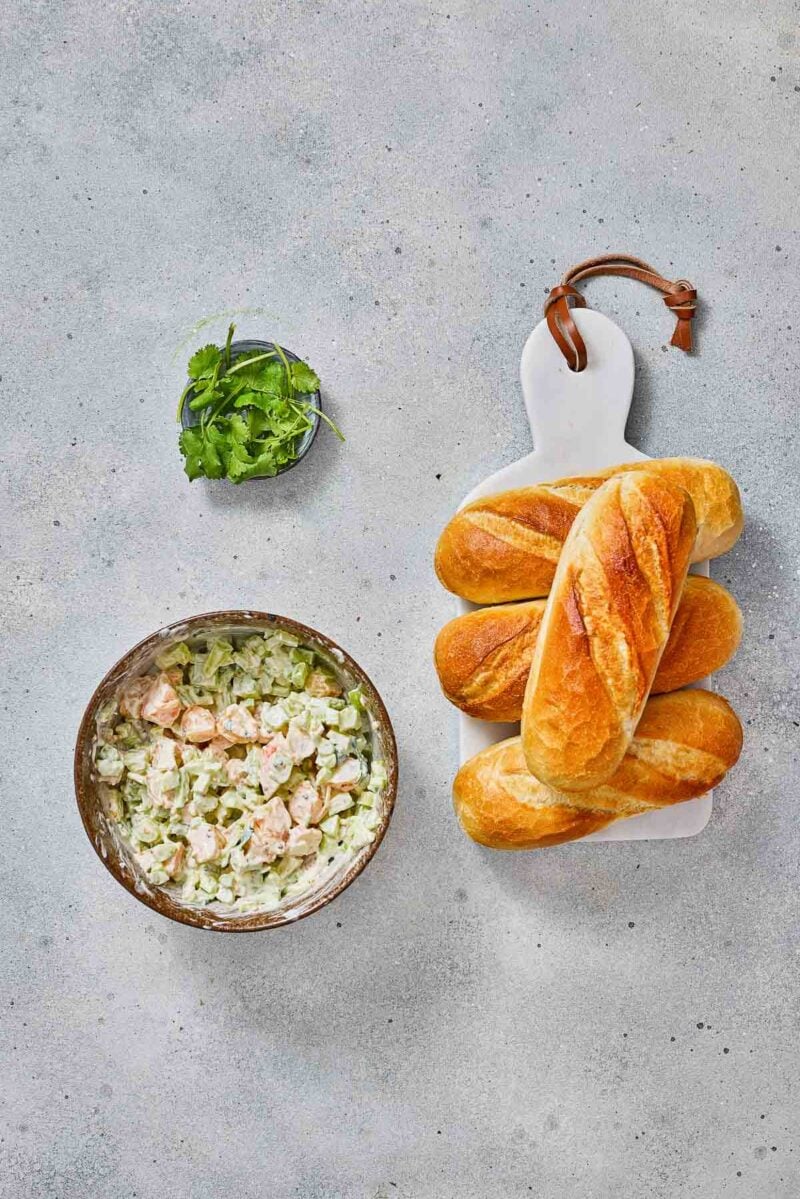 A bowl of shrimp salad next to a bunch of sandwich rolls on a white cutting board, with a small dish of fresh herbs above.