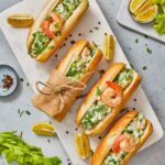 A marble board with four shrimp rolls garnished with herbs, surrounded by lemon wedges, spices, and celery on a light gray surface.
