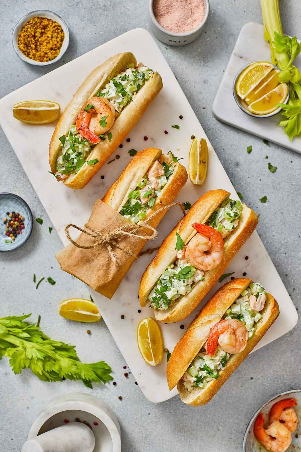 A marble board with four shrimp rolls garnished with herbs, surrounded by lemon wedges, spices, and celery on a light gray surface.