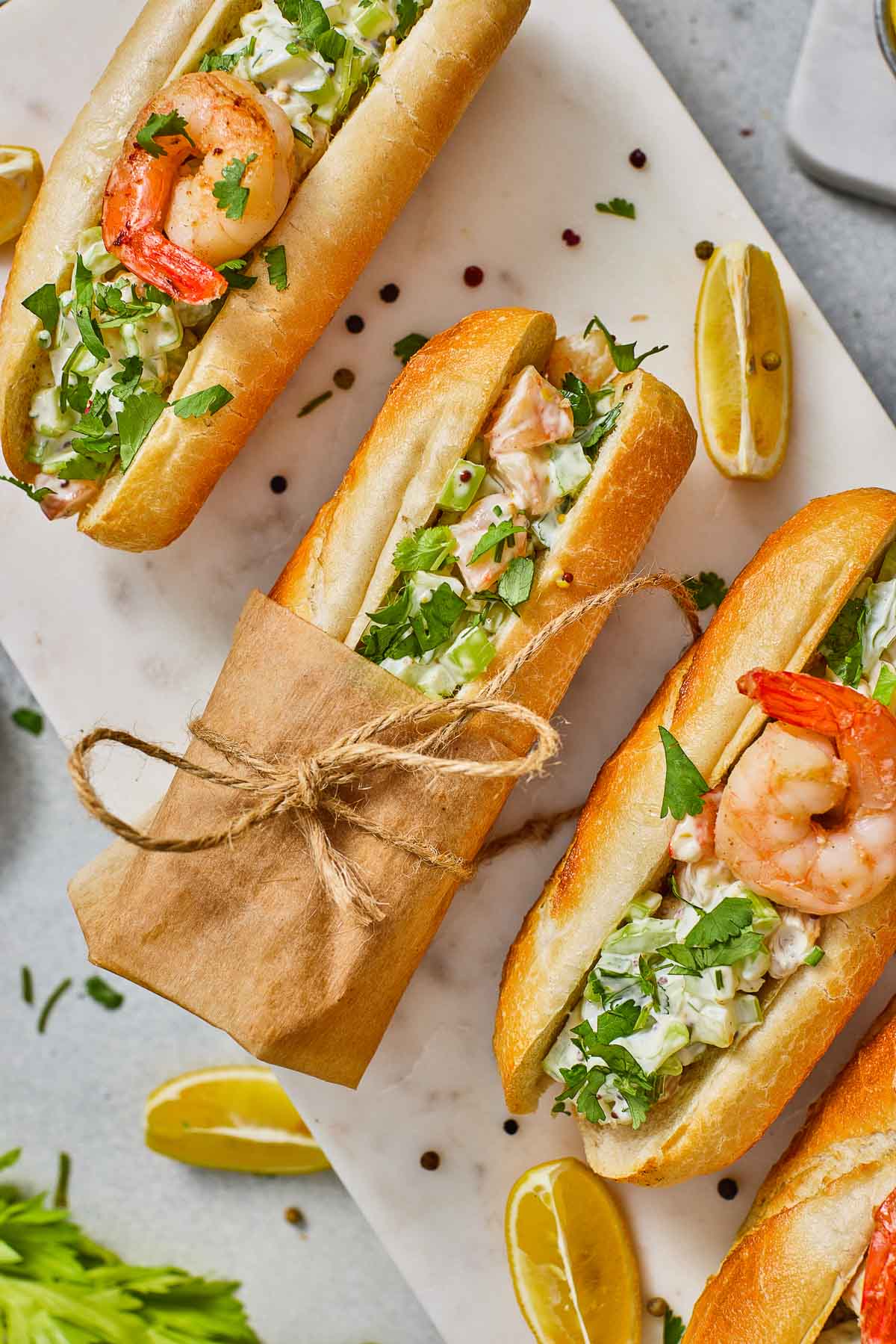 Three shrimp rolls with lettuce and herbs in toasted buns, garnished with lemon wedges, on a white surface; one roll is wrapped in brown paper and tied with string.