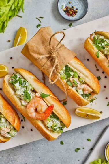 Four east coast shrimp rolls on baguettes are arranged on a marble surface, one wrapped in brown paper and tied with twine, garnished with lemon wedges and herbs.