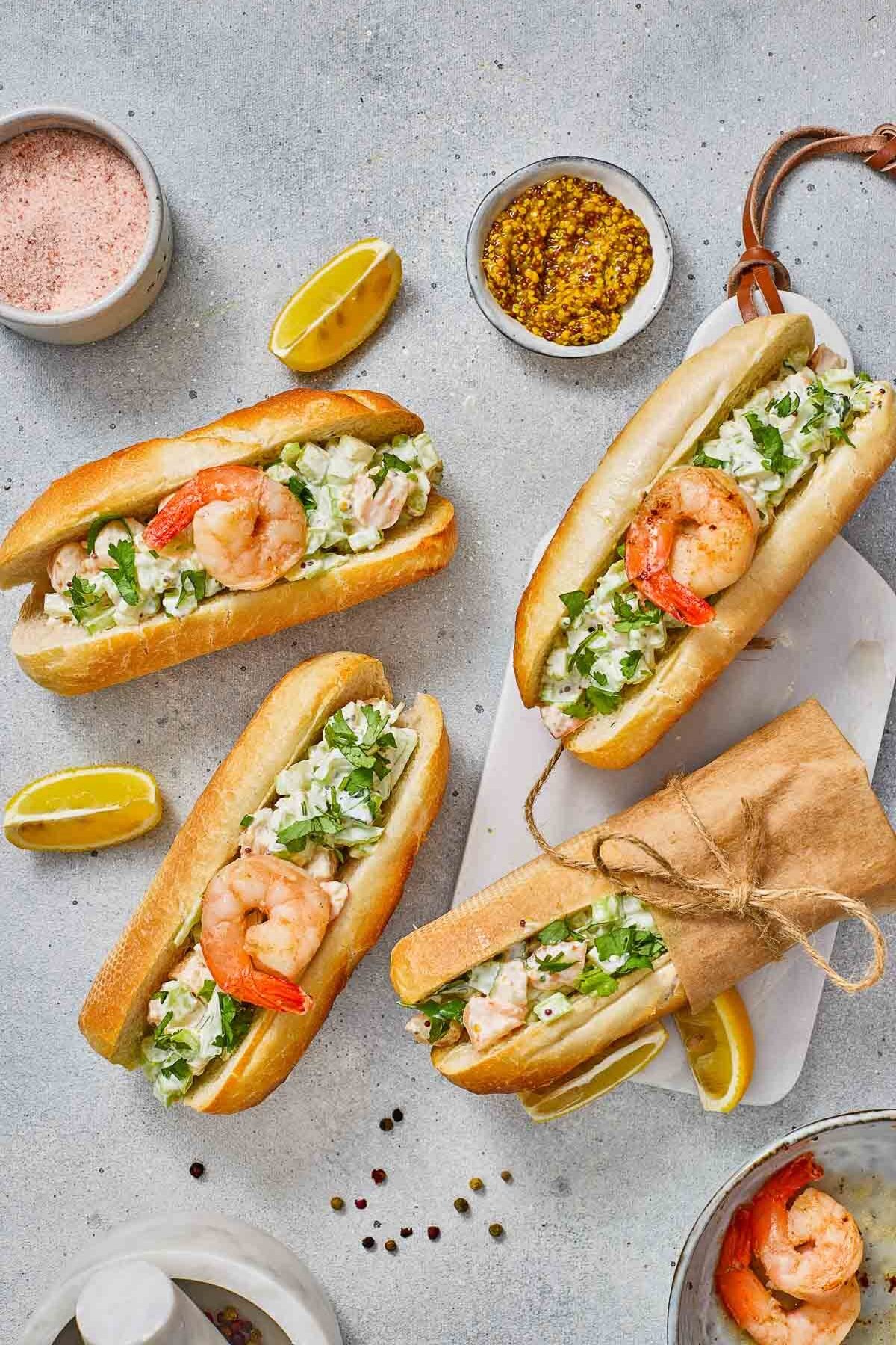 Shrimp rolls with fresh herbs and a shrimp on top in baguettes are arranged on a light surface, surrounded by lemon wedges, mustard, and seasoning bowls.